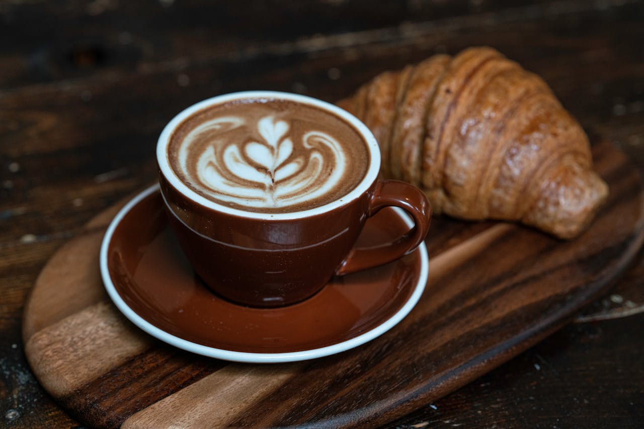 about-us A close-up of a latte with beautiful art served alongside a croissant on a rustic wooden board.
