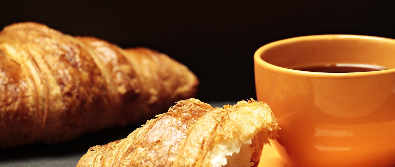 heros-img Close-up of a freshly baked croissant paired with a warm cup of coffee on a table.